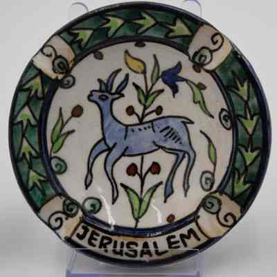 One of the four painted ceramic ashtrays given to the Jordans by a ruler in Bombay.: Up close image of one of the four painted ceramic ashtrays. This one is painted with a white background with a blue antelope in the middle. It surrounded by green leaves and blue swirls. Around the outside there are green leaves and it reads "JERUSALEM" across the bottom in black text.