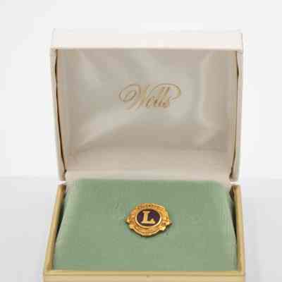 The gold and blue Lions Club pin in a white and gold Wells box.: Image of the gold and blue Lions Club pin in mint green felt in a white "Wells" box with gold trim. "Wells" is written in cursive on the inside top cover of the box.