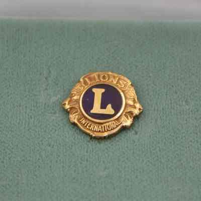 The gold and blue Lions Club pin in a white and gold Wells box.: An up close image of the gold and blue Lions Club pin on a mint green felt. The pin is roughly half an inch in size and reads "LIONS INTERNATIONAL" with a large "L" in the middle. There are also two lion heads on either side of the "L."