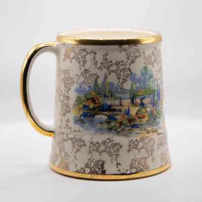 Mug-shaped music box: The image shows a ceramic mug-shaped music box adorned with gold details. It features an intricate scene depicting a stream, surrounded by a landscape, and the text  "in an old world garden."