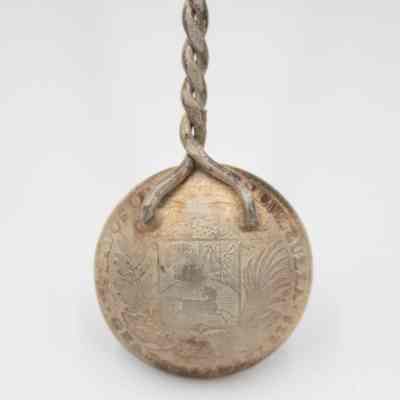 Back of 1932 Bolivar Spoon: Closeup of just the back of spoons bowl. At the top, the twisted rope stem is soldered on to the bowl. Around the top edge it says "Estados Unidos De Venezuela." The coin features the Coat of Arms of Venezuela with a white horse, a bundle of wheat and weaponry that includes a sword, a sabre and three lances.