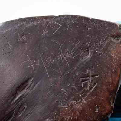 Bottom of carved wooden figurine of a seated woman.: Up close image of the bottom of the carved wooden figurine. It is etched and engraved with multiple Chinese characters.