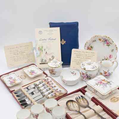 Full set of the Royal Crown Derby items received by Mrs. Jessie Jordan from various events at the Kuwait Masonic Lodge 6810.: Image of the full set of Royal Crown Derby items received by Mrs. Jessie Jordan. There is a blue Masonic Presentation Bible propped up in the back along with three invitations from various Ladies' Nights. All of the porcelain items are white and have the same floral pattern decorating them. There is one larger bowl, a cream pitcher, two sugar dishes (one large, one small), a smaller bowl, two cigarette ashtrays, a cigarette box, a set of six silver knives in a red case, a pair of silver salad serving spoons in a red case, and six small teacups.