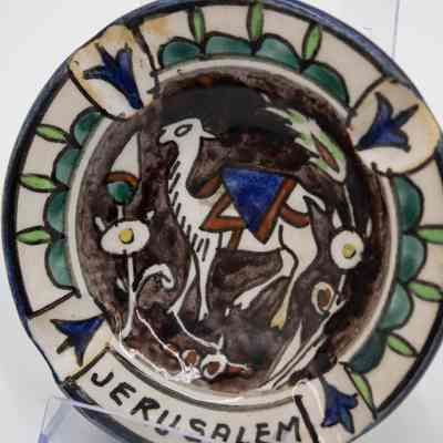 One of the four painted ceramic ashtrays given to the Jordans by a ruler in Bombay.: Up close image of one of the four painted ceramic ashtrays. This one is painted with a purple background with a white camel in the middle wearing a blue and brown saddle blanket. Around the outside there are two colors of green designs and it reads "JERUSALEM" across the bottom in black text.