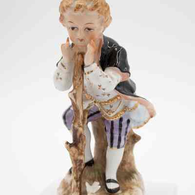 Boy painted porcelain figurine.: Image of one of two hand-painted porcelain figurines depicting a boy dressed in romantic-era fashion. He is holding a stick and playing with a small white bird, likely a dove.