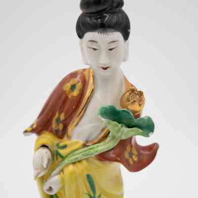 Guanyin porcelain figurine.: Up close image of the painted white porcelain figurine dressed in a yellow robe and red shawl. The figurine is holding a gold lotus blossom.