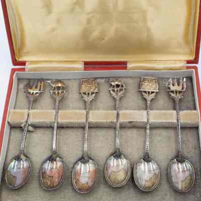 Set of six coffee spoons paired in sets of two with three designs.: Up close image of the set of six silver coffee spoons with two depicting Arabian camels, two with Arabic script, and two with abstract Kuwaiti dhows. They are resting on beige felt in a red case and yellow satin lid.