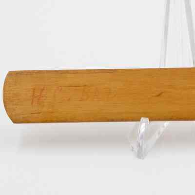 Writing on mini cricket bat: A miniature wooden cricket bat rests on a clear stand, displaying handwritten text, "H.C. BAT."