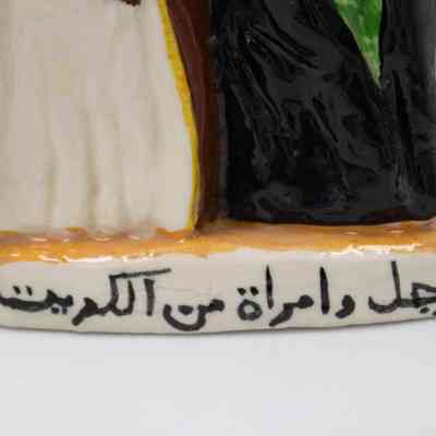 Base of figure with Arabic script.: A ceramic figurine depicts a man and woman in traditional Kuwaiti attire, standing together. Underneath there is black Arabic script that translates into "A man and woman from Kuwait."