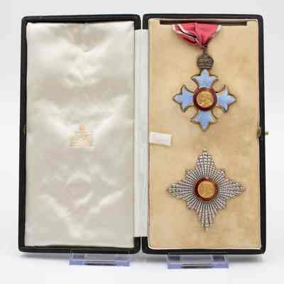 Open box with Knight of the British Empire medal: The image shows an open box displaying a Commander of the British Empire medal, featuring a cross with blue enamel and a central gold medallion, accompanied by a star-shaped Knight of the British Empire medal beneath it.