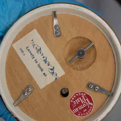 Bottom of music box: The bottom of the music box has screws, a winding key and two sticker labels. One has a mountain range, and the words, "Lovely thing to look at." The red sticker says "Product of Yeoman Plate, England."