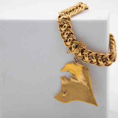 Charm bracelet given to Mrs. Jessie Jordan by His Highness Sheikh Ahmad Al Jaber Al Sabah.: Up close image of the heavy gold bracelet with one charm of Kuwait draped over a white foam block. The charm shows the location of Ahmadi, denoted by a small ruby and star. The chain on the bracelet is a thick gold rope.