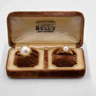 Pair of gold and pearl rings in a "Bell's San Antonio" double ring box.: Image of double brown felt ring box with a pair of gold and pearl rings. Each of the rings sit on a brown felt pillow and the inside of the box is yellow satin. The inside lid reads "TEXAS OLDEST JEWELRY STORE BELL'S SAN ANTONIO" in black letters. The ring on the left has a larger pearl, prong setting, and a narrow band, while the one on the right has a smaller pearl with a wide, tapered band.