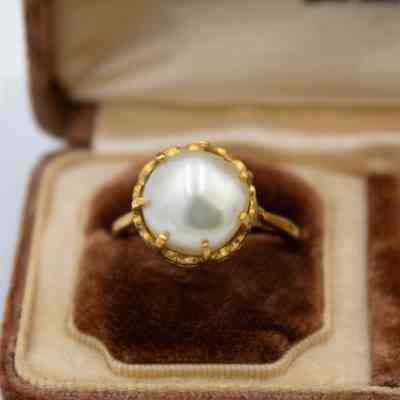 Gold and pearl ring with a larger pearl, prong setting, and a narrow band on a brown felt pillow.: Up close image of the left gold and pearl ring on a brown felt pillow. The ring has a large sphere-shaped pearl set in a seven-prong setting and a narrow gold band.