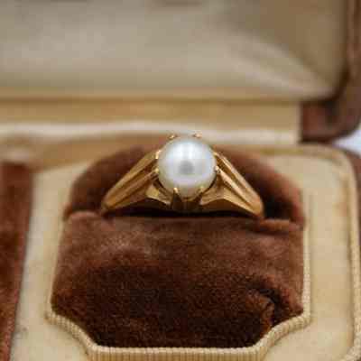 Gold and pearl ring with a smaller pearl with a wide, tapered band on a brown felt pillow.: Up close image of the right gold and pearl ring on a brown felt pillow. The ring has a wide, tapered band and a sphere-shaped pearl in a gold four-prong setting.