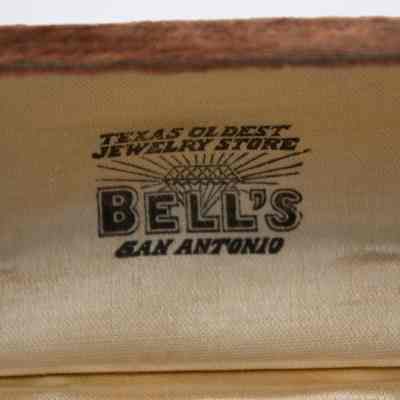 Inside of the double ring box.: Up close image of the yellow satin interior of the lid of the double ring box. Black writing reads "TEXAS OLDEST JEWELRY STORE BELL'S SAN ANTONIO."