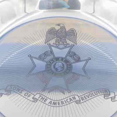 The engraving on the sterling silver Sons of the American Revolution plate.: Up close image of the engraving on the center of the silver Sons of the American Revolution plate. The engraving reads "SONS OF THE AMERICAN REVOLUTION" across the bottom on a ribbon. There is an eagle sitting on a four-point cross and a seal that reads "LIBERTAS ET PATRIA" with the profile head of George Washington in the middle.