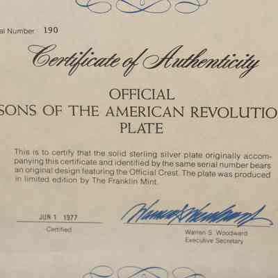 Certificate of Authenticity for the Sons of the American Revolution Silver Plate.: Up close image of the Certificate of Authenticity. It is a cream-colored document with black text: "Serial Number 190," "Certificate of Authenticity," "OFFICIAL SONS OF THE AMERICAN REVOLUTION PLATE," "This is to certify that the solid sterling silver plate originally accompanying this certificate and identified by the same serial number bears an original design featuring the Official Crest. The plate was produced in limited edition by The Franklin Mint." It is also "Certified Jun 1 1977" and signed by "Warren S. Woodward Executive Secretary" in blue ink.