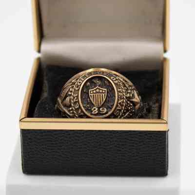 L.T. Jordan's 1929 class ring from Texas A&M University.: Up close image of L.T.'s class ring in its case. L.T.'s class ring is gold with eagle and shield on the front. There is an embossed "29" under the shield, representing his class year. Around the center it reads "A&M COLLEGE OF TEXAS" The ring sits on a black pad inside a black box with gold trim. The ring is in what is known as the "antique" style, with black coloring in the grooves of the gold.
