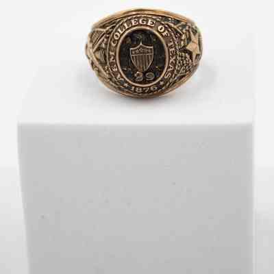 Image of the front of L.T. Jordan's 1929 class ring from Texas A&M University.: Up close image of L.T.'s class ring sitting on a white block. L.T.'s class ring is gold with eagle and shield on the front. There is an embossed "29" under the shield, representing his class year. Around the center it reads "A&M COLLEGE OF TEXAS." The ring is in what is known as the "antique" style, with black coloring in the grooves of the gold.