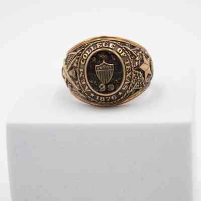 Image of the front of L.T. Jordan's 1929 class ring from Texas A&M University.: Up close image of L.T.'s class ring sitting on a white block. L.T.'s class ring is gold with eagle and shield on the front. There is an embossed "29" under the shield, representing his class year. Around the center it reads "A&M COLLEGE OF TEXAS." The ring is in what is known as the "antique" style, with black coloring in the grooves of the gold.