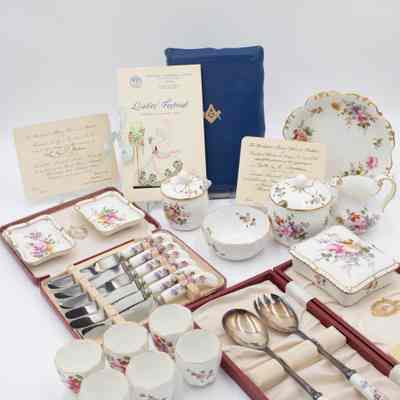 Complete collection of Royal Crown Derby porcelain given to Mrs. Jessie Jordan by the Masonic Lodges in Kuwait.: Image of the full set of Royal Crown Derby items received by Mrs. Jessie Jordan. There is a blue Masonic Presentation Bible propped up in the back along with three invitations from various Ladies' Nights. All of the porcelain items are white and have the same floral pattern decorating them. There is one larger bowl, a cream pitcher, two sugar dishes (one large, one small), a smaller bowl, two cigarette ashtrays, a cigarette box, a set of six silver knives in a red case, a pair of silver salad serving spoons in a red case, and six small teacups.