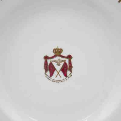 Royal seal on Royal Crown Derby China plate.: Up close image of the royal seal in the center of the plate. The plate is white and the seal is gold and red. The seal has a crown on the top with a gold falcon and two crossed red flags. The flags read "KUWAIT" and the text on the bottom says "J.A.J.AL-SABAH" in both English and Arabic