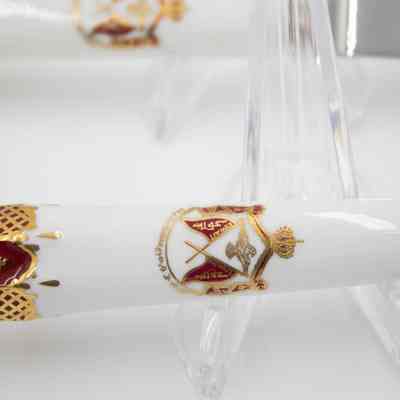 Handle of the Royal Crown Derby fork.: Up close image of the white porcelain handle decorated with the Kuwaiti royal seal. The seal has a crown on the top with a gold falcon and two crossed red flags. The flags read "KUWAIT" and the text on the bottom says "J.A.J.AL-SABAH" in both English and Arabic