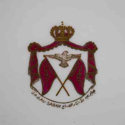 Royal seal on Royal Crown Derby China plate.: Up close image of the royal seal in the center of the plate. The plate is white and the seal is gold and red. The seal has a crown on the top with a gold falcon and two crossed red flags. The flags read "KUWAIT" and the text on the bottom says "J.A.J.L-SABAH" in both English and Arabic