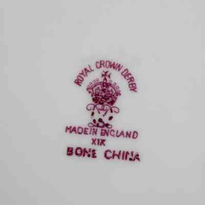 Makers mark on the back of the Royal Crown Derby China plate.: Up close image of the maroon maker's mark on the back of the white China plate. The mark is stamped with an English crown with "ROYAL CROWN DERBY" and "MADE IN ENGLAND." There is also the number "XIX" and "BONE CHINA" clarified under the location specification.