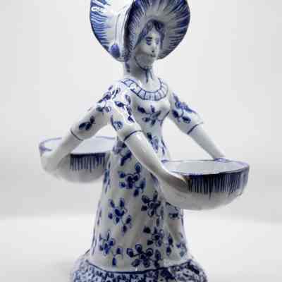 Blue and White Two-Sided Figurine Woman Holding Bowls: A white porcelain doll with blue details on her dress and bonnet. She holds a bowl out with both hands. The backside of the doll is identical to the front, having a second doll connected to the first along her back.