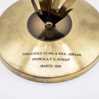Brass Flower Clock Base: Close-up image of the base of the Brass Flower Clock. The brass is slightly dirty and tarnished. At the front of the base is an enscription in black capital letters which reads "Presented to Mr. & Mrs. Jordan From H.A.F.C. Kuwait March 1961."
