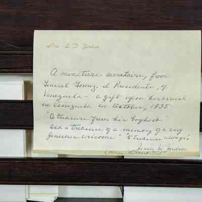 Note inside table: A handwritten note describes a miniature folding desk gift from 1935, set against its wooden backdrop. Mrs. L.T. Jordan writes, "a minature secretaire, from General Gomez, el Presidente, of Venezuela- a gift upon arrival in Venezuela in October, 1935. "A treasure from his [illegible]" and a Treasure of a memory of a [illegible] welcome. "A treasure always." Jessie W. Jordan (Mrs. Leland T.)"