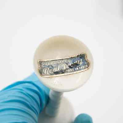 Bottom of the carved ivory Chinese puzzle ball with pedestal.: Up close image of the bottom of the carved ivory pedestal. There is a black sticker with gold writing. It reads "Guaranteed Real Ivory Made in India."