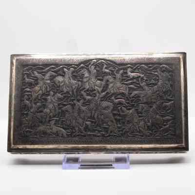 Top of the small chased silver box debossed with a hunting scene.: Image of the rectangular silver box debossed with a hunting scene. It depicts a monabat-kari-style decoration of a hunting scene of Bahram V, with figures on horseback pursuing various animals with dogs. There are lions, rabbits, foxes and dear, all within a rectangular reserve and arched pediment. The edge is bordered with birds in foliage (golo morph) and guilloche below.