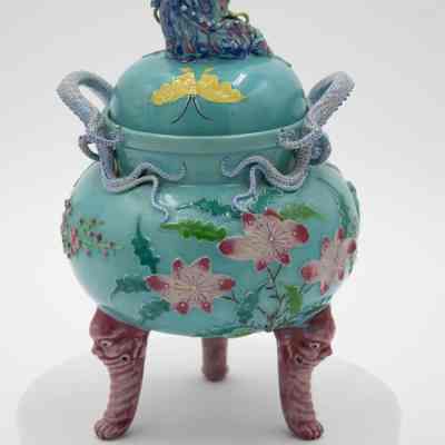 Back of the turquoise Chinese footed incense burner from Hong Kong.: Image of the back of the turquoise incense burner decorated with pink lotus flowers, blue and green leaves, and a yellow moth. The burner has three legs that are pink with faces.