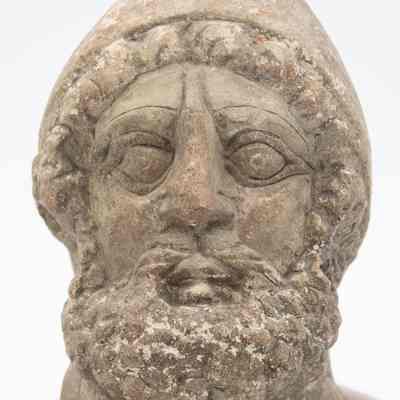 Close up Roman stone head: Close up on the Roman man's head carved out of stone. He has curly hair and a beard, with a pointed helmet covering most of his hair. His eyebrows are thin, his eyes are wide, and his nose has visible wear.