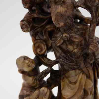Carved stone scene of a woman and child on a ram.: Up close image of the carved stone scene of a woman surrounded by flowers next to a small child riding a ram. The woman is holding a lotus flower and has a rounded hat on her back. The piece is marbled brown.