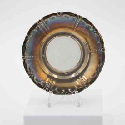 Saucer: Image of a single silver-plated saucer on a plastic stand. The middle divot of the saucer remains porcelain while the rest of the surface is plated with silver. The silver is heavily tarnished.