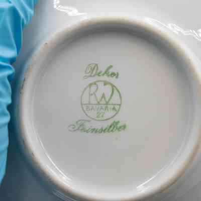 Makers Mark: Close-up image of a saucer's bottom.  The bottom is porcelain. Printed with green ink, there is a circle with an "R W" lettermark, as well as "Bavaria 27" inside the circle. Outside it it is surrounded with cursive that reads "Dekor Feinsilber".