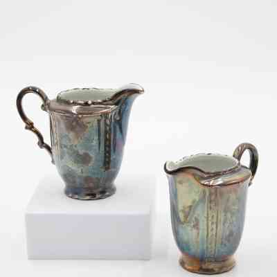 Creamers: Image of a pair of silver-plated creamers with handles on each. The silver on them is heavily tarnished.