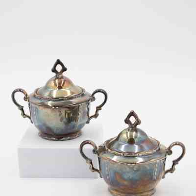 Creamers: Image of a pair of silver-plated handled creamers with lids. The silver on them is heavily tarnished.