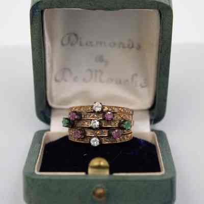 The 5-banded Harem ring given to Mrs. Jessie Jordan by Mr. Conrad Hilton.: Up close image of the 5-banded Harem ring. The Harem ring is made of 5 bands attached with a bar and set with gemstones. There are three white gems, four purple, and two green. It is sitting on a dark blue felt pillow inside a mint green ring box.