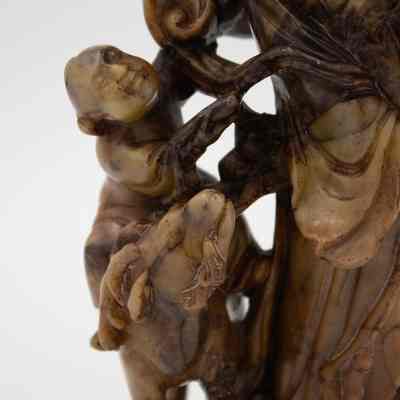 Carved small child riding a ram.: Up close image of the small child looking up at the woman and riding on a ram. It is marbled brown.