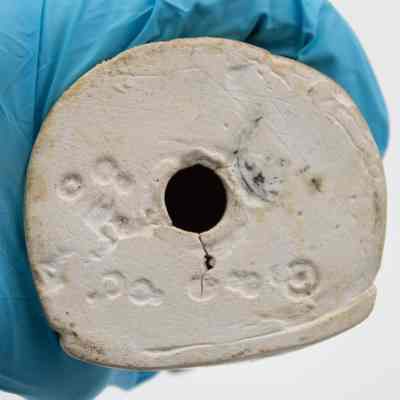 Bottom of figurine.: Image of the white ceramic bottom of the figurine. There is a large hole, used when firing the piece in the kiln. There are also nine imprinted circles and the remnants of a black stamped seal.