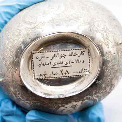 Bottom of the Persian silver bowl.: Up close image of the bottom of the Persian silver bowl. The bottom has a white sticker with black text that reads: "Fadavi Jewelry - Silver and Gold Factory of Isfahan Mithqal 28 Abar 84."