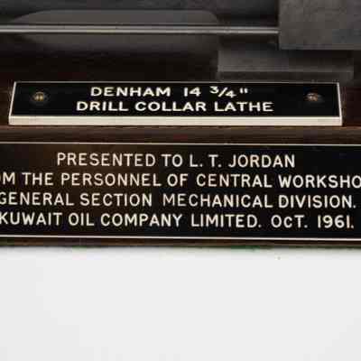 The two plaques located on the miniature drill collar lathe.: Up close image of the two plaques on the miniature drill collar lathe. The plaque on the top reads "DENHAM 14 3/4 DRILL COLLAR LATHE," while the one on the front reads "PRESENTED TO L.T. JORDAN FROM THE PERSONNEL OF CENTRAL WORKSHOPS GENERAL SECTION MECHANICAL DIVSION. KUWAIT OIL COMPANY LIMITED. OCT. 1961."The plaques are black with white writing on attached to the dark wood base.