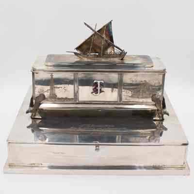 Silver casket humidor given to Mr. L.T. Jordan when he retired from the Kuwait Oil Company in 1962.: Image of the silver casket humidor on silver pedestal. On top is a Kuwaiti dhow with LTJ initials enameled on the front. The casket humidor depicts scenes of Kuwaiti oil operations with a miniature dhow on the top and falcons as the legs. Additionally, the box containing the book with signatures is engraved with “L.T. Jordan K.B.E. from his friends and colleagues in the Kuwait Oil Company Ltd. 1948-1961.”