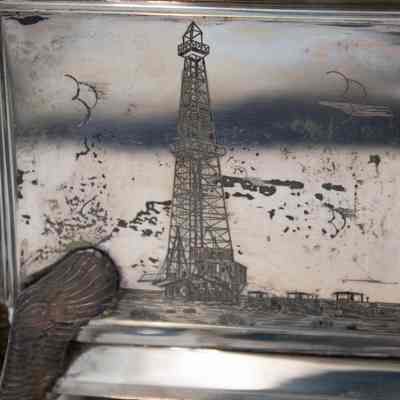 Engraved design on the front of the silver casket humidor.: Up close image of the engraved decoration on the front of the silver casket humidor. It shows an oil rig set up.