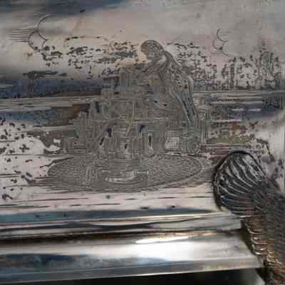 Engraved design on the front of the silver casket humidor.: Up close image of the engraved decoration on the front of the silver casket humidor. It shows a man working on a oil rig.